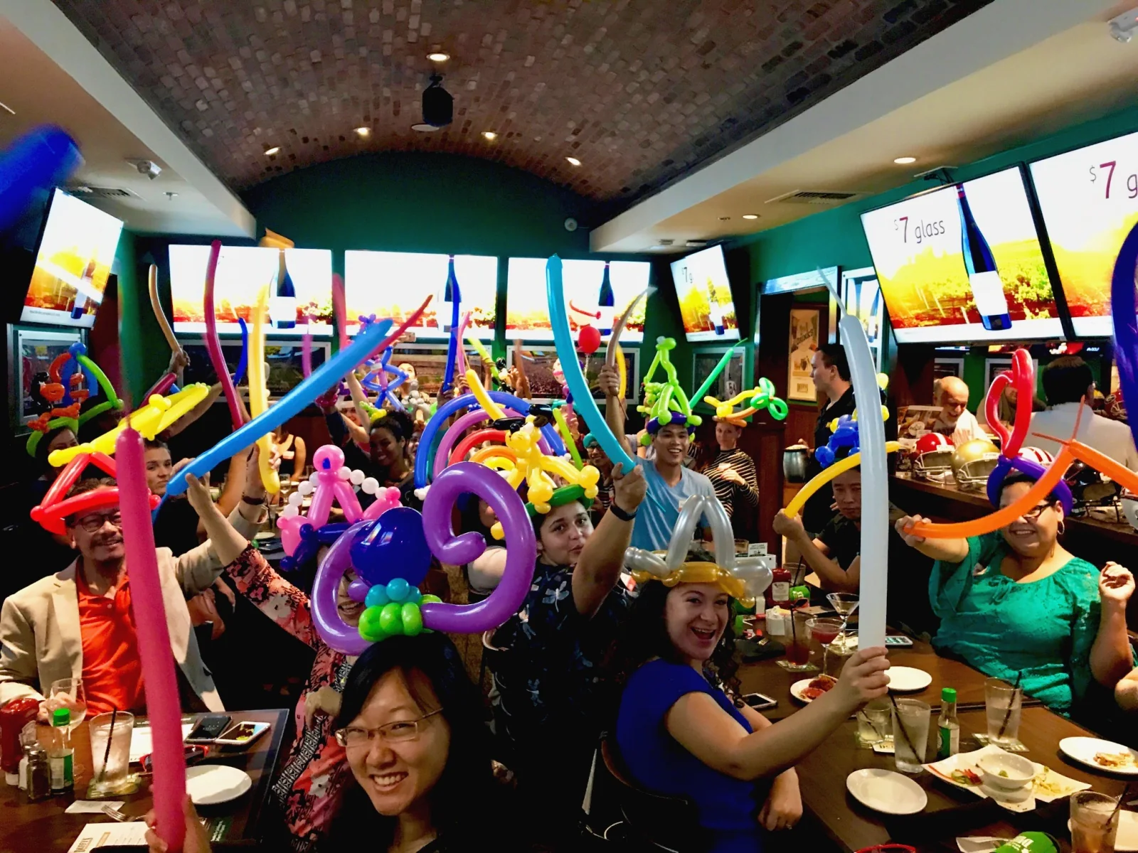 A large group of adult restaurant guests raising colorful balloon creations.